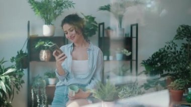 Video of smiling woman drinking a cup of coffee while using her mobile phone in greenhouse.