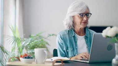 Video of beautiful senior woman working with laptop while checking some documents in the kitchen at home. 