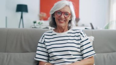 Video of kind senior woman in glasses resting on sofa at home.