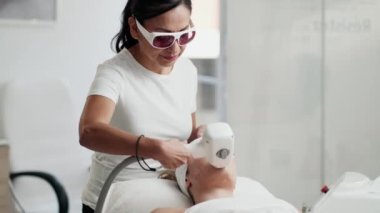 Terapist kozmetik uzmanının videosu kaplıca güzellik kliniğinde kadının yüzüne lazer tedavisi uyguluyor..