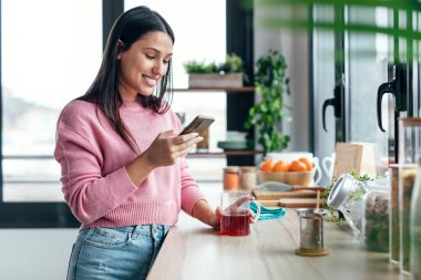 Shot of beautiful woman using her mobile phone while drinking a cup of tea in the kitchen at home.