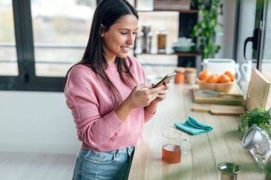 Shot of beautiful woman using her mobile phone while drinking a cup of tea in the kitchen at home.