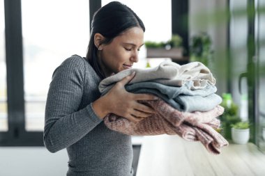 Shot of beautiful young woman holding and smelling clean clothes at home.