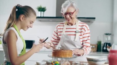 Video of cute granddaughter taking photos with her smartphone while grandmother making tasty chocolate cake in the kitchen.