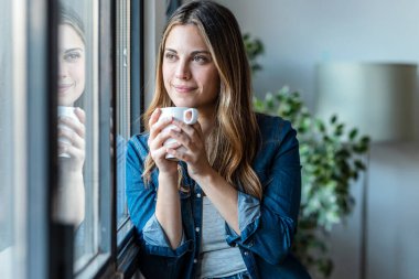 Evdeki pencerenin yanında beklerken bir fincan kahve içen güzel genç bir kadın..