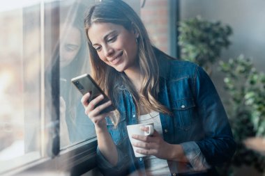 Evdeki oturma odasının penceresinin yanında kahve içerken güzel bir genç kadının cep telefonunu kullanması.