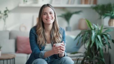 Evdeki oturma odasında kanepede otururken bir fincan kahve içen güzel genç bir kadının videosu..