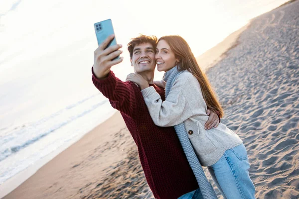 拍到一对年轻貌美的夫妇在一个寒冷的冬天在海滩用手机拍照 — 图库照片