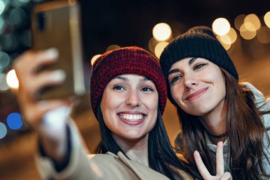 Gece şehirde barış jesti gösterirken iki neşeli kadının akıllı telefonuyla selfie çekmesi.