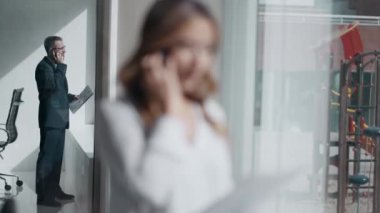 İş arkadaşı modern bir ofiste arka planda cep telefonu kullanırken akıllı telefonlu çekici bir kadının pencerenin önünde konuşma videosu..