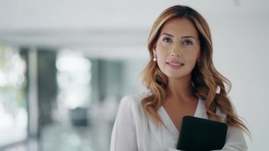 Modern bir ofiste kameraya bakarken elinde dijital tablet olan çekici ve zeki bir iş kadını videosu..
