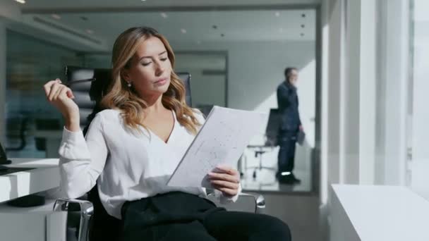 現代のオフィスで窓の前に座っているいくつかの文書を分析魅力的なビジネス女性のビデオ — ストック動画
