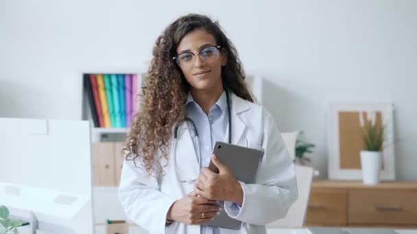 病院の診察室に立っているカメラを見ながらデジタルタブレットを持っている美しい若い医師の女性のビデオ — ストック動画