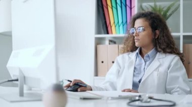 Hastanenin konsültasyonunda bazı notlar alırken bilgisayardaki hastayla video görüşmesi yapan güzel genç doktor kadının videosu.