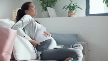 Evde, oturma odasındaki kanepede dizüstü bilgisayarla çalışan, karnı ağrıyan endişeli hamile kadının videosu.