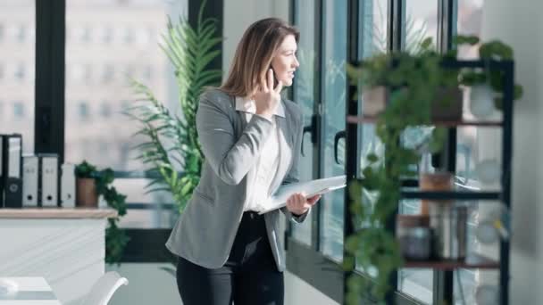 在一间现代化的办公室里 一位漂亮优雅的女商人站在窗边 一边用智能手机聊天一边工作的视频 — 图库视频影像