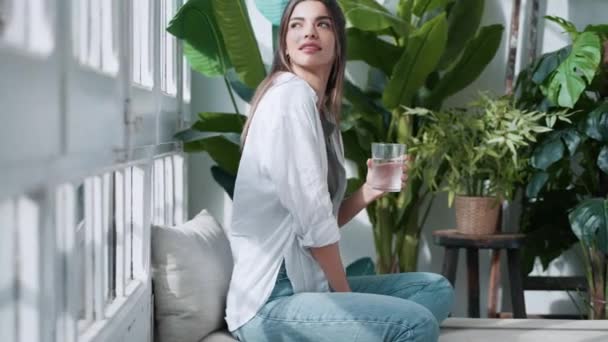 自宅で植物でいっぱいのリビングルームでソファに座っている間にハスの位置飲料水で若い女性のビデオ — ストック動画