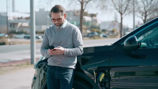 電気ガソリンスタンドで電気自動車を充電しながら携帯電話を使用してエレガントな男のビデオ — ストック動画