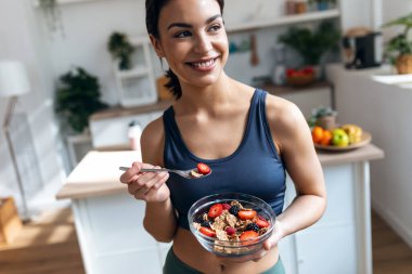 Shot of athletic woman eating a healthy bowl of muesli with fruit in the kitchen at home clipart