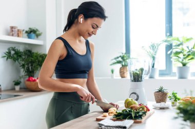 Shoot of athletic woman cutting fruits and vegetables to prepare a smoothie while listening to music with earphones in the kitchen at home clipart