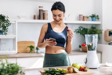 Evdeki mutfakta smoothie hazırlarken atletik bir kadının cep telefonu kullanması..