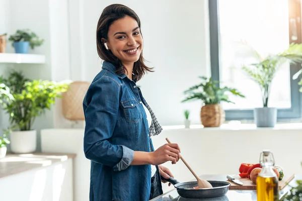 부엌에서 이어폰으로 음악을 아름다운 미소짓는 여자가 야채를 요리하는 — 스톡 사진