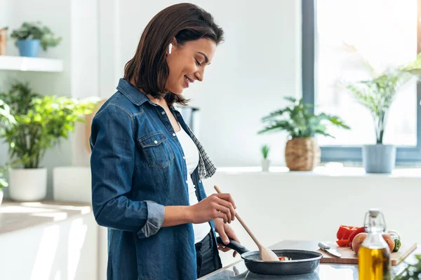 부엌에서 이어폰으로 음악을 아름다운 미소짓는 여자가 야채를 요리하는 — 스톡 사진