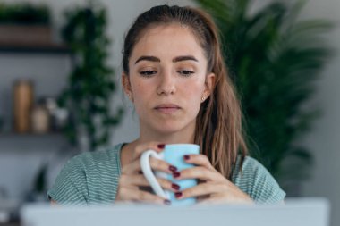 Evdeki mutfakta bir fincan kahve içerken dizüstü bilgisayarla çalışan güzel bir kadının fotoğrafı..