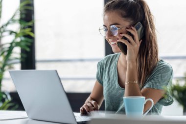 Evdeki mutfakta akıllı telefonla konuşurken dizüstü bilgisayarla çalışan gülümseyen bir iş kadını..