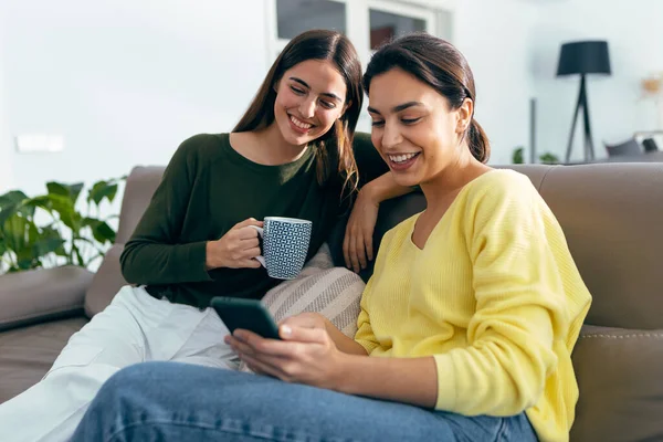 自宅のリビングルームでソファに座っているスマートフォンを見ている間に話している2人の笑顔の若い女性のショット — ストック写真