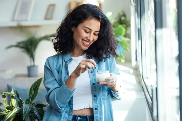 stock image Shot of happy beautiful woman eating yogurt while standing in living room at home.