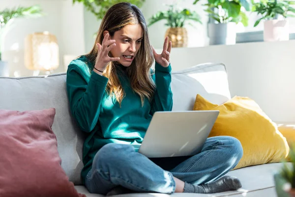 Shot of angry frowning young woman working online on laptop sitting on couch at home