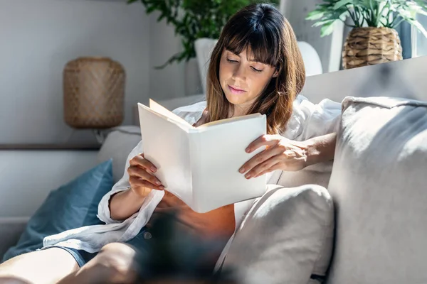 家でソファに座っている間に本を読んでいるかなり若い女性のショット — ストック写真