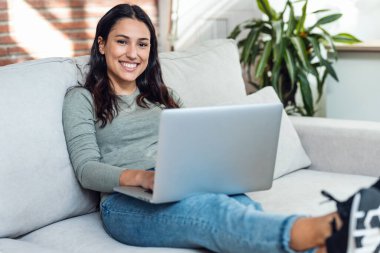 Evdeki kanepede otururken dizüstü bilgisayarıyla çalışan güzel bir genç kadının fotoğrafı..