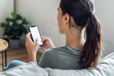 Evdeki kanepede oturan genç bir kadının cep telefonu kullanışının görüntüsü.