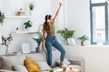 Evde kulaklıkla müzik dinlerken, koltukta şarkı söyleyip dans eden mutlu bir kadın..