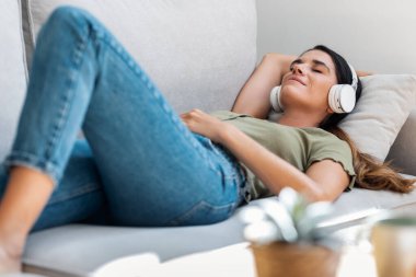 Evde kanepede uzanmış kulaklıkla müzik dinleyen güzel bir kadın.
