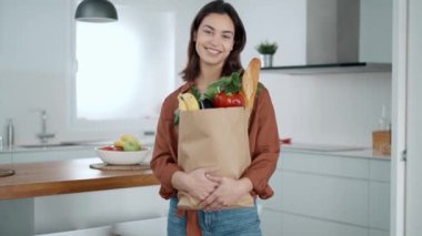 Evdeki mutfakta kameraya bakarken elinde sebze dolu bir poşet tutan güzel bir genç kadının videosu. 