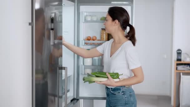 自宅のキッチンで冷蔵庫にいくつかの健康的な食べ物を保っている美しい若い女性のビデオ — ストック動画