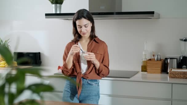 自宅のキッチンで先を探しながらヨーグルトを食べている美しい幸せな女性のビデオ — ストック動画