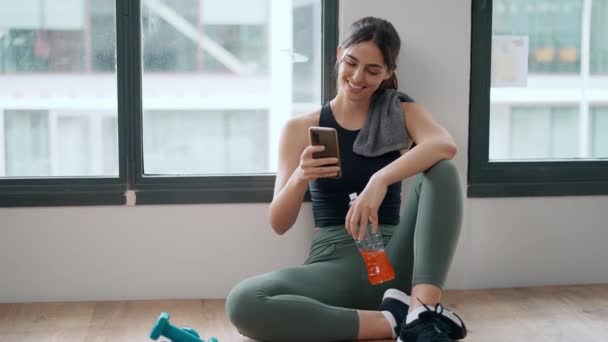 スポーティな若い女性のビデオ 彼女の携帯電話を使用して 自宅でジムの後でタンパク質の揺れを保持しながら — ストック動画