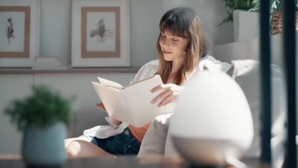 ビデオのかなり若いです女性読みますA本に座っている間ソファに家 — ストック動画