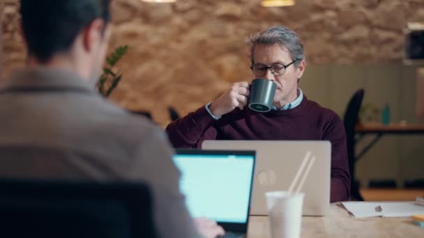 成熟英俊的企业家在同事间与笔记本电脑一起工作时喝一杯咖啡的视频 — 图库视频影像