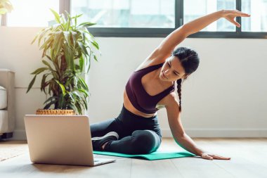 Spor yapan genç bir kadının evdeki dizüstü bilgisayarla online beden eğitimi derslerini takip ederek yoga egzersizleri yapması..