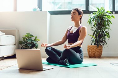 Spor yapan genç bir kadının evdeki dizüstü bilgisayarla online beden eğitimi derslerini takip ederek yoga egzersizleri yapması..