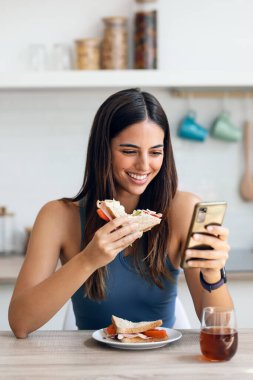 Güzel, sportif bir kadının evde akıllı telefon kullanırken sağlıklı sanviç yediği bir fotoğraf.