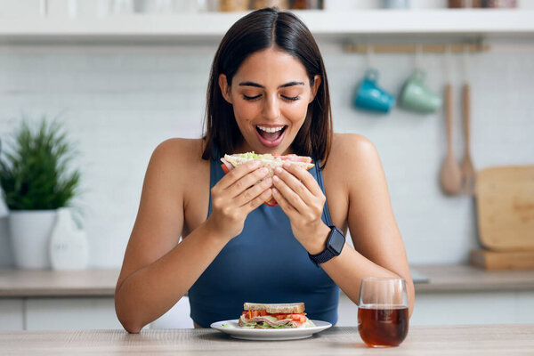 Снимок красивой спортивной женщины едят здоровый сандвич, глядя на камеру на кухне дома