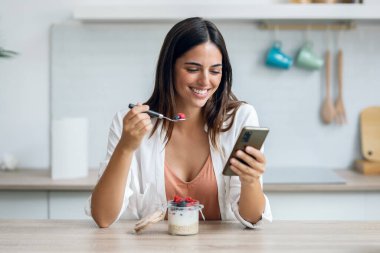 Shot of beautiful woman eating a porridge with berries while using smartphone in the kitchen at home clipart