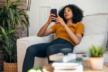 Evdeki kanepede otururken gülümseyen bir kadının cep telefonunu kullanması..