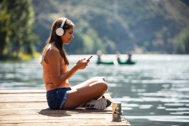 Güzel bir kadının kulaklıkla müzik dinlerken gölün yanında akıllı telefon kullanması.